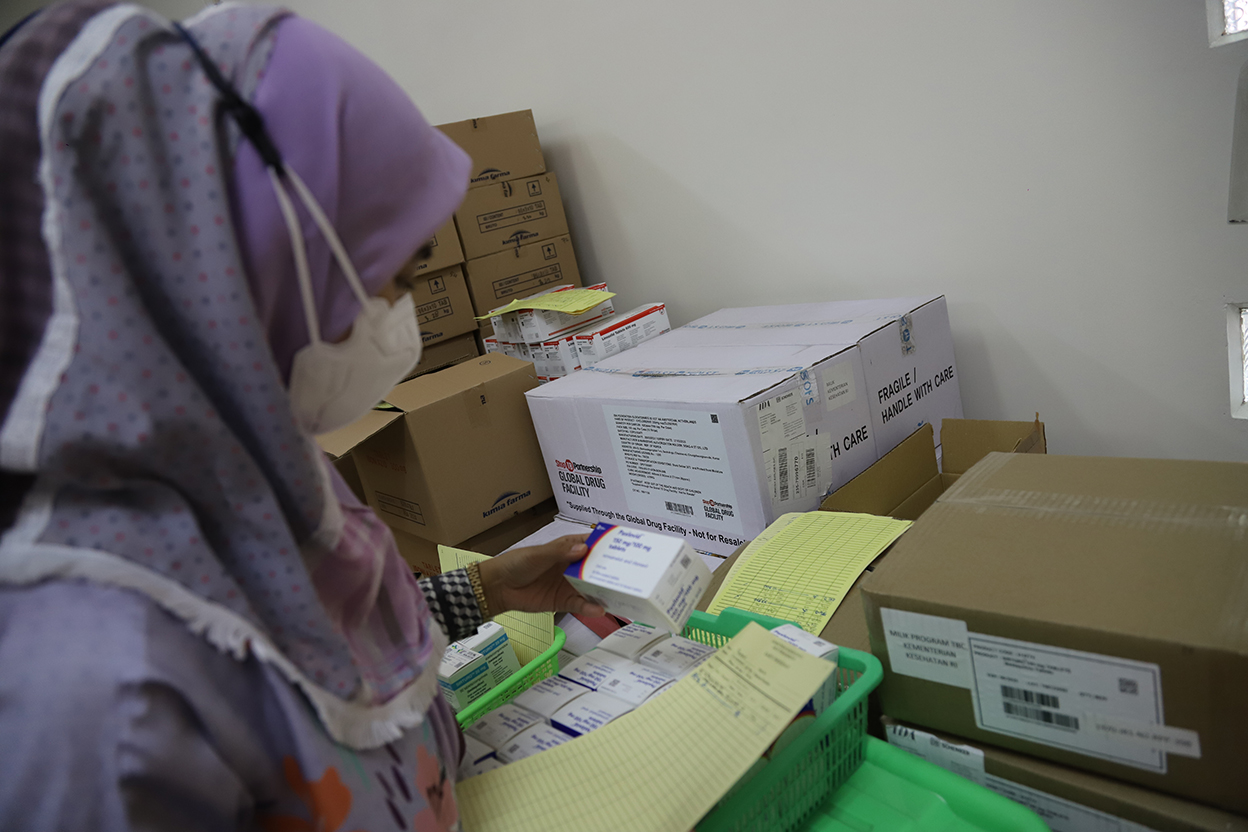 IMG_8822_ID The image shows an individual in a hijab examining stacked boxes of medication. They are holding and looking at a package of medicine, seemingly conducting inventory or stock checking in a storage room or pharmacy.