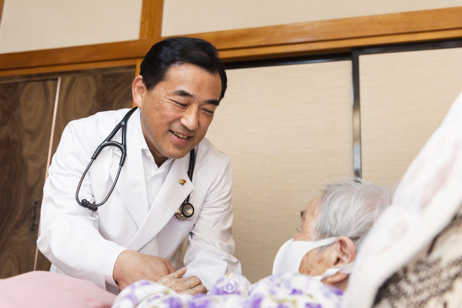 A doctor bends down to meet with an older patient in her hospital bed in Japan