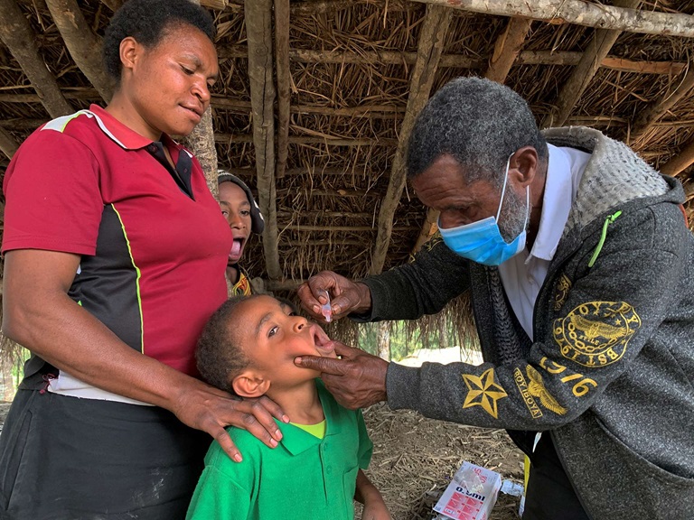Polio A health worker holds a child's chin and dips their head back to administer a polio vaccine. The child's mother rest her hands on his shoulders.